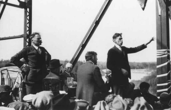 Black-and-white photograph of men speaking and listening on a raised platform, with a seated audience in the foreground and a rural landscape behind them.