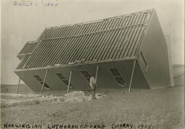 1908 photo of a church tipped upside-down with a man standing in front of it. Text on the photo says, Norwegian Lutheran Church Dovray 1908. Built 1898