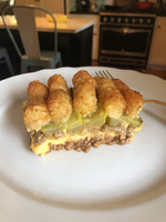 A serving of tater tot hotdish (showing its layers of ground meat, cheese, pickles, and tater tots) sits atop a white plate, with a kitchen scene in the background.