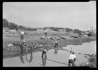 Black and white photograph of a construction site along a creek. Six workers lay wood, stone, and stakes in the creek, while two onlookers watch from the land next to the water.