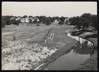 Lightly sepia-toned black and white photograph of Ȟaȟá Wakpádaŋ (Bassett Creek) curving through a field with a baseball diamond. White houses and tall, dark trees line the background.