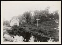 Black-and-white photograph of a spring at Bassett Creek in the foreground and a sign that reads “Inglewood Park and Springs.” Behind the creek is a white wooden house and a grassy hill with trees and a trail.