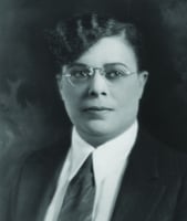 The head and shoulders of a woman with permed, side-swept bangs, wearing a suit and tie.