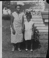 An African American woman and her young daughter stand in front of a house splattered with paint.