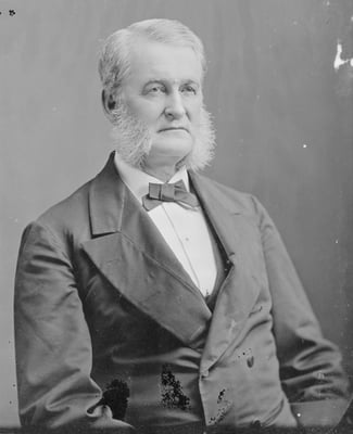 Seated man wearing a suit and bow tie with white hair, including white mutton-chop whiskers.