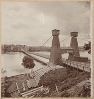 Sepia photograph of a suspension bridge. Stacks of logs are piled up in the foreground.