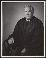A man in a black robe sits next to a chair, with his right hand resting on it.