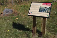 A sign with text and a color image stands on two wooden supports in front of a grassy background. A historical marker set into the earth is visible at left.