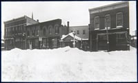 Four buildings line a wintery street scene, with snow piled up in the foreground.