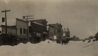 Piles of snow line a street with buildings lined up on the left and right sides, with telephone poles marking both.