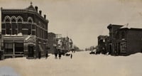 A snowy corner in a street, with buildings on either side and people in the center.