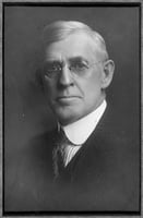 The head and shoulders of a man wearing a chin-height stiff collar, with white hair and glasses.
