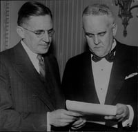 The torsos, heads, and hands of two men wearing ties. Both men are holding and looking at a piece of paper.