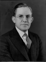 Head and shoulders of a man wearing a suit and tie with a collar and glasses.