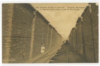 Railroad tracks run through an alley created by two rows of stacks wooden boards at the left and right, rising up to a sepia-colored sky.