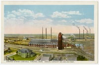 Bird's-eye view of a lumber mill showing a red multistory building with a red silo next to it, smaller buildings in the foreground, and smokestacks in the background, all under a blue sky with white clouds.