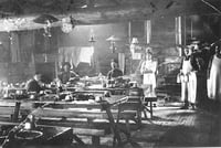 Three people sit and three others, wearing aprons, stand inside a dining hall with tables, benches, and an exposed wooden rafter overhead.
