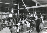 The interior of a lumber camp building showing three long tables with laborers sitting on benches. The building's rafters are visible above them.