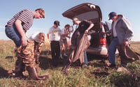 Six people stand grouped together in front of a minivan with its trunk door open. Three of the people prepared to put on waders..
