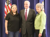 Three people stand in front of a blue curtain with the American and Minnesota state flags.