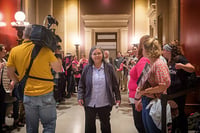 A woman in a blue button-up shirt and cardigan walks through a hallway with lobbyists surrounding her and a cameraman at the left of the image.
