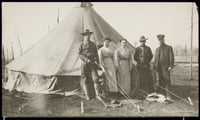 Three men (two of them in military uniform) and two women post in front of a round canvas tent, with the charred remains of trees to the right and left.