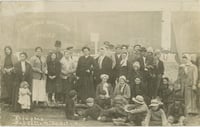 A crowd of people stands in front of a Canadian Northern train car. Women and children cluster in the front, with men closer to the back. They wear hats, kerchiefs, shawls, and coats.