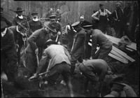 Men in military uniforms holding shovels stand around a circle of earth, with planks of wood to their right and more people and forest behind them.