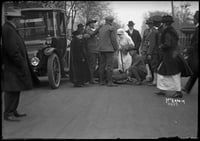A man watches from the left foreground (an automobile stands behind him) watching a group of aid workers attending to a fire victim or victims.