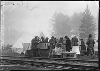 Behind a train track and in front of a stand of trees, workers distribute food and drinks to men in uniforms.