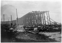 Railroad tracks above a trestle rise to the right and then stop abruptly where they've been burned. Fire damage is all around them.
