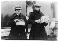 Two women wearing caps and long coats hold loaves of bread and other food rations in their hands as refugees disembark from a train behind them.