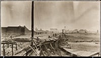 A destroyed structure sits in the foreground next to a standing wooden pole, with charred buildings and fire survivors in the background.