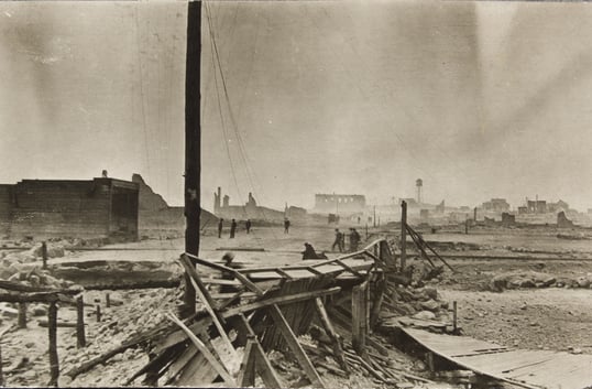 A destroyed structure sits in the foreground next to a standing wooden pole, with charred buildings and fire survivors in the background.