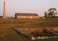 Landscape at sunset with foundation ruins in the foreground and a building and upright pillar in the background.