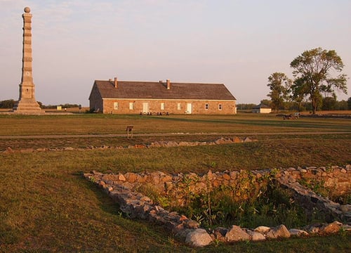 Landscape at sunset with foundation ruins in the foreground and a building and upright pillar in the background.