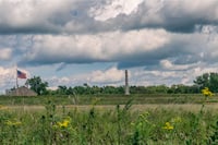 Grassy foreground with yellow flowers below a blue but cloud-filled sky, with a flag at left and a pillar at center.