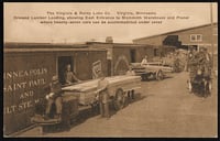 A wagon loaded with lumber sits in the middle ground, with workers preparing to load it into a one-story warehouse on the left. More forehouses are visible in the background.