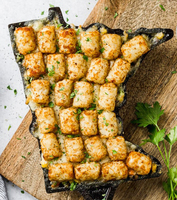 A baking dish shaped like the state of Minnesota filled with browned tater tots, with filling visible below them, against a cutting-board background.