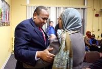 A man in a blue suit jacket holds the left arm of a woman wearing hijab, with yellow walls and other people behind them.
