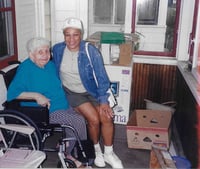 A woman in a wheelchair sits next to a woman in a blue denim jacket who perches next to her. They are inside an indoor porch.