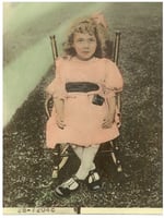 A little girl with a formal dress, hair bow, and Mary J ane shoes sits in a chair in an outdoor setting.The black-and-white photo has been colorized so that the girl's dress and hair ribbon are pink. 