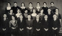 Students stand in three staggered rows of seven people each. They wear formal dresses and suits.