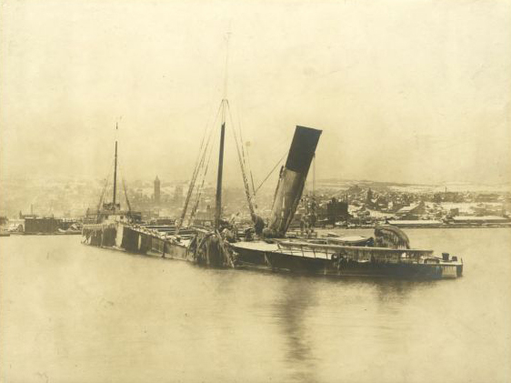 In a sepia-tinted photograph, a steamship with a central smoke funnel sits in water beside a harbor broken in half.