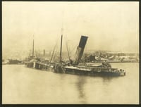 A wrecked steamship sits partially submerged in water, with its bow facing the shore of a lake, with buildings behind it.