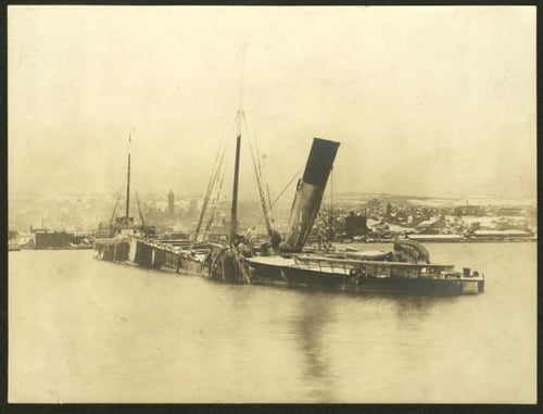 A wrecked steamship sits partially submerged in water, with its bow facing the shore of a lake, with buildings behind it.