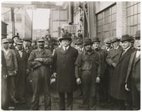 Men wearing caps and brimmed hats pose in front of factory buildings.