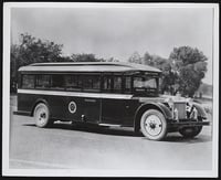 1920s-era bus viewed at a shallow three-quarter angle in front of a tree line with sky in the background