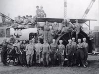 Women workers waring hats and overalls pose in front of a tractor.