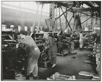 Two women in overalls stand in the left foreground, working with machines; two other women workers stand at right in the middle ground. Line shafts branch out above their heads.